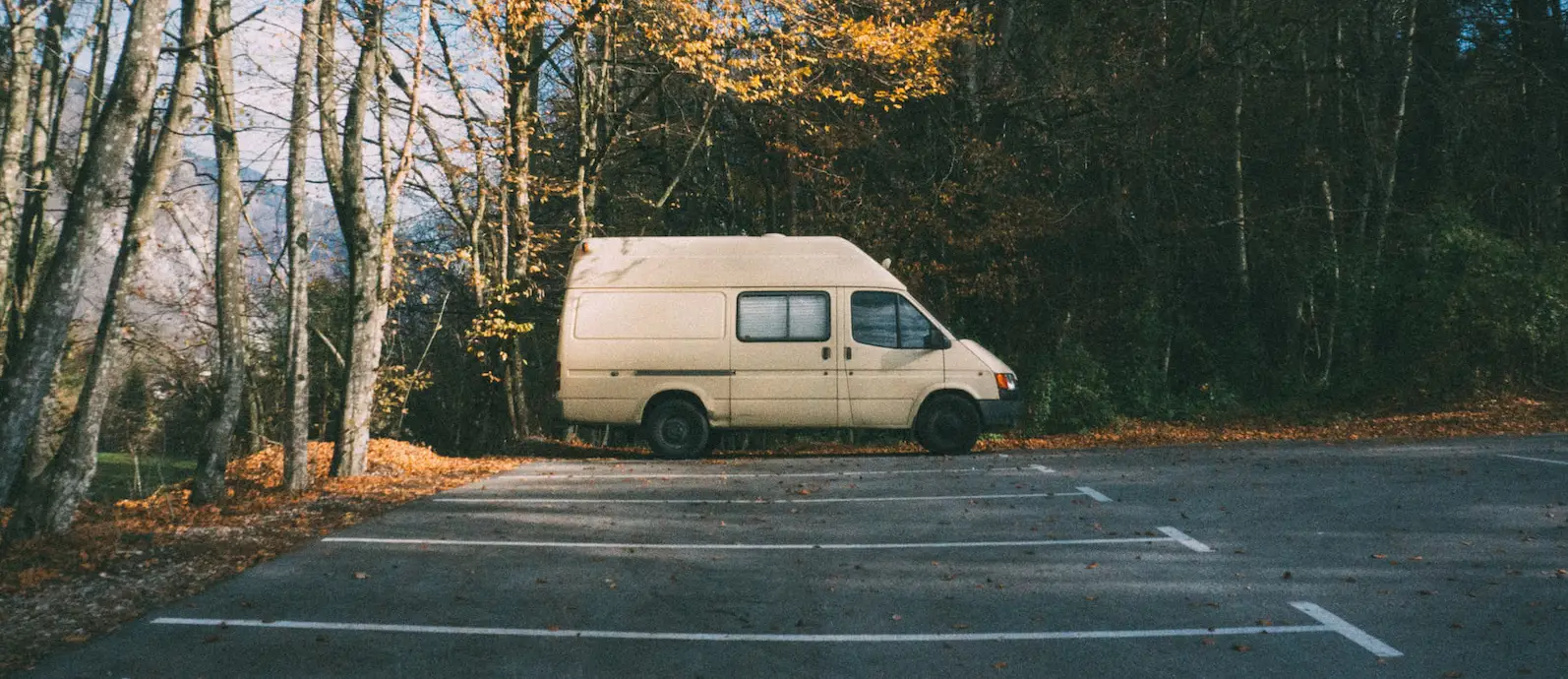 side view of a white van parked in an empty car park surrounded by autumnal trees